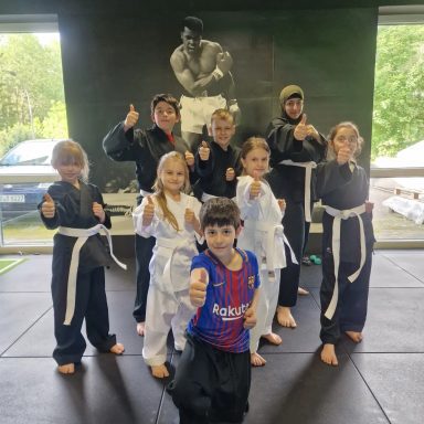 Junge und Mädchen in Kampfsportanzügen posieren mit Daumen hoch, im Hintergrund ein Schwarz-Weiß-Bild.